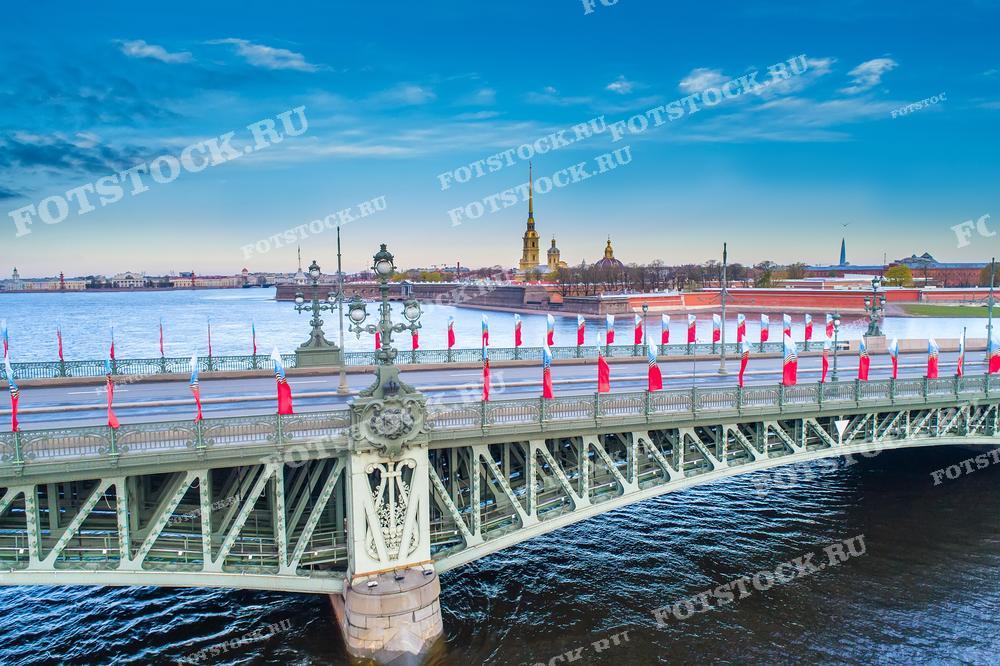 Этот снимок показывает один из мостов Санкт-Петербурга — Троицкий. Мост украшен флагами, что добавляет цвета и праздничности обычно строгому городскому ландшафту. На фоне видны силуэты Петропавловской крепости и её золочёные шпили. Фотография может быть использована в туристических брошюрах, публикациях о знаменитых мостах и архитектурных символах города, о праздновании Дня Победы в Петербурге.